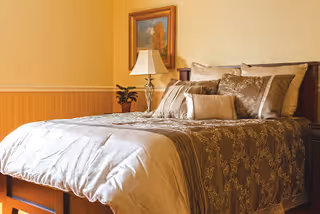 A neatly made bed with multiple decorative pillows and a patterned bedspread in a warmly lit bedroom. A bedside table with a lamp and a small plant is visible, along with a framed painting on the wall.