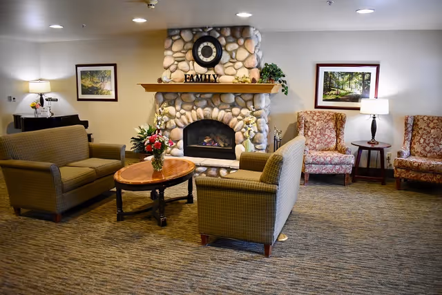 Seating area in a senior living common room with sofas and armchairs facing a stone fireplace topped with a clock and a 'FAMILY' sign.