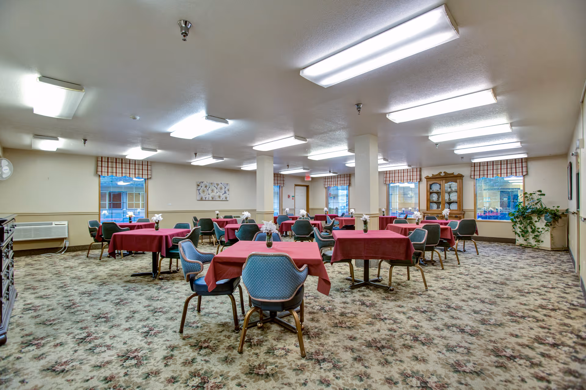 Spacious senior dining room with multiple tables covered in red tablecloths and chairs on patterned carpet.