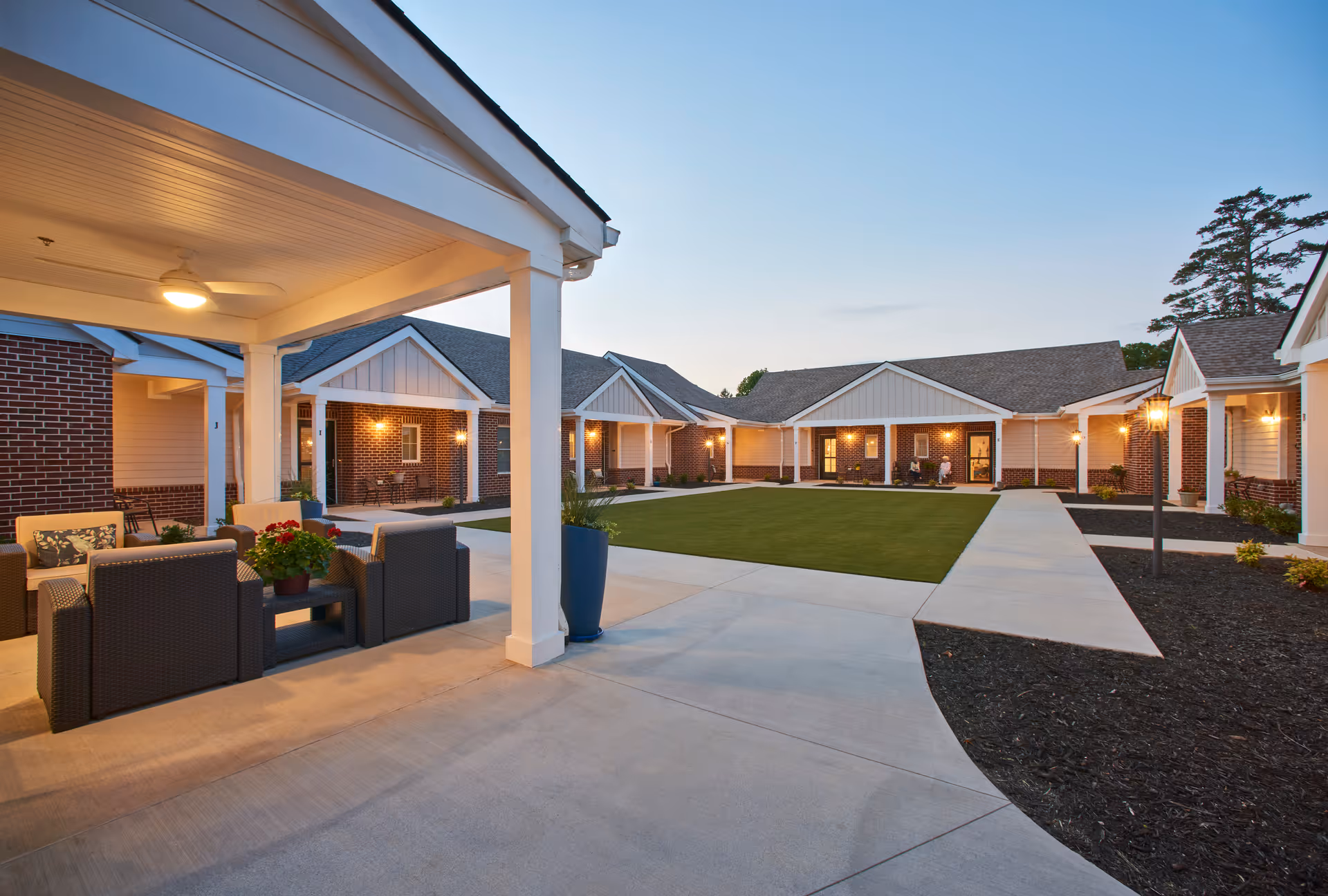 Outdoor courtyard area of a senior living facility with covered seating area featuring cushioned chairs and a table with potted plants. The courtyard is surrounded by single-story buildings with brick and white siding, lit by warm exterior lights during dusk.
