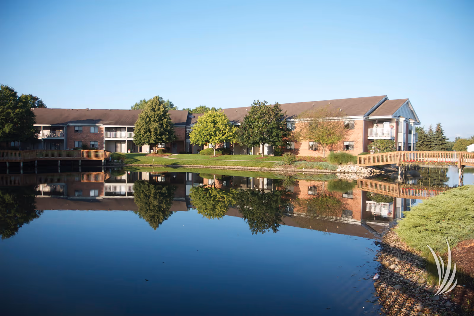 A two-story brick building with balconies and a wooden bridge over a calm pond reflecting the building and surrounding trees, under a clear blue sky.