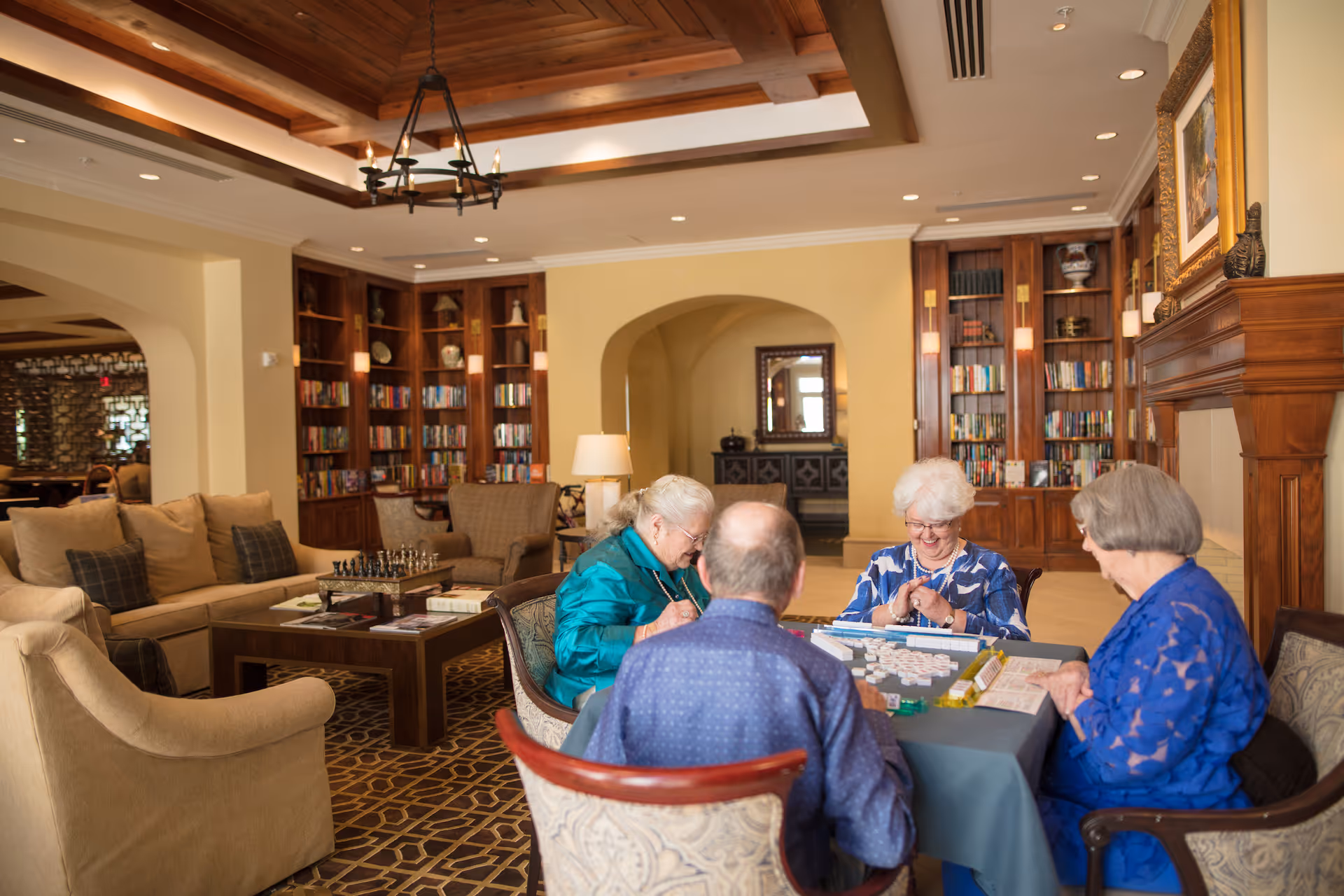 Four elderly people sitting around a table playing a tile-based game in a cozy, well-lit living room with wooden bookshelves, a fireplace, and comfortable seating.