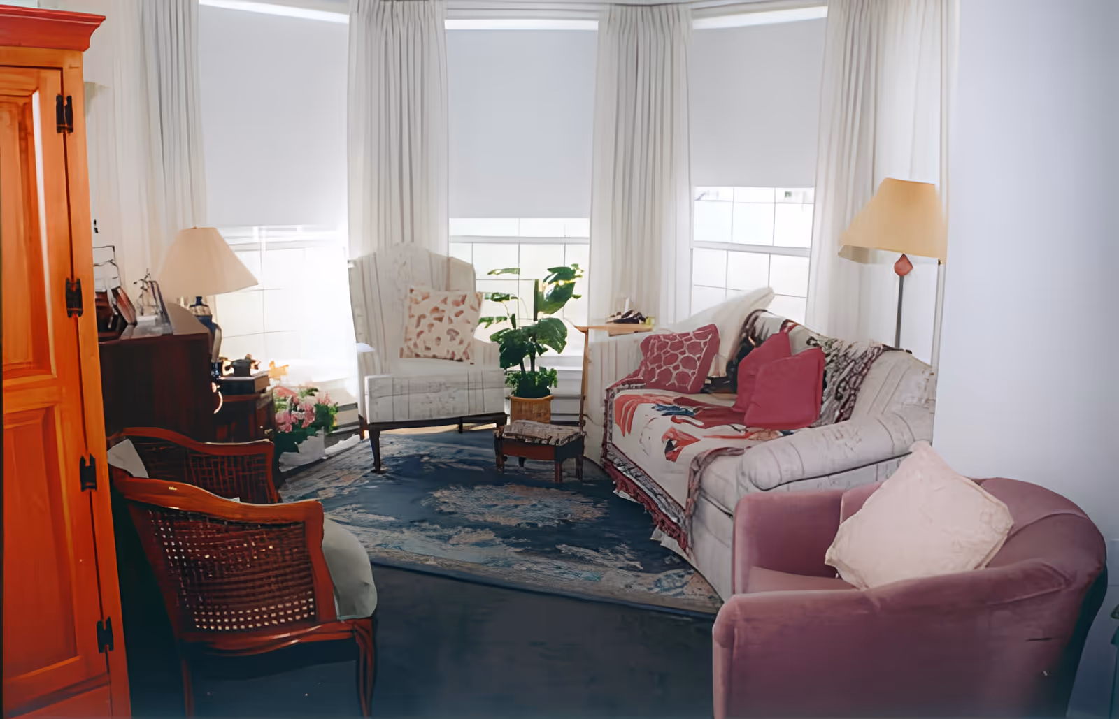 A cozy living room with a white couch adorned with red and patterned pillows, a purple armchair with a light pillow, a white armchair with a patterned pillow, a wooden chair with a woven back, a wooden cabinet, two lamps, a small wooden table, a plant, and large windows with white curtains and blinds letting in natural light.