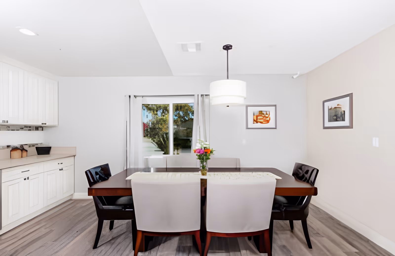Dining area with a rectangular wooden table surrounded by chairs, a hanging pendant light, and white cabinetry along the left wall.
