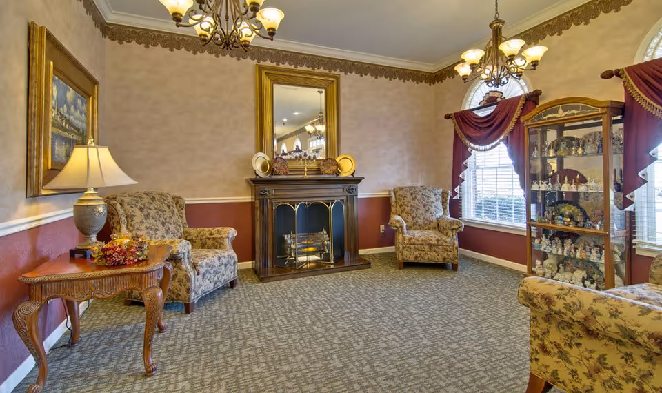 A cozy senior living room with floral upholstered armchairs, a wooden side table with a lamp and decorative items, a fireplace with a large mirror above it, and a glass display cabinet filled with figurines. The room has patterned carpet, beige and maroon walls with decorative trim, and windows with red drapes.