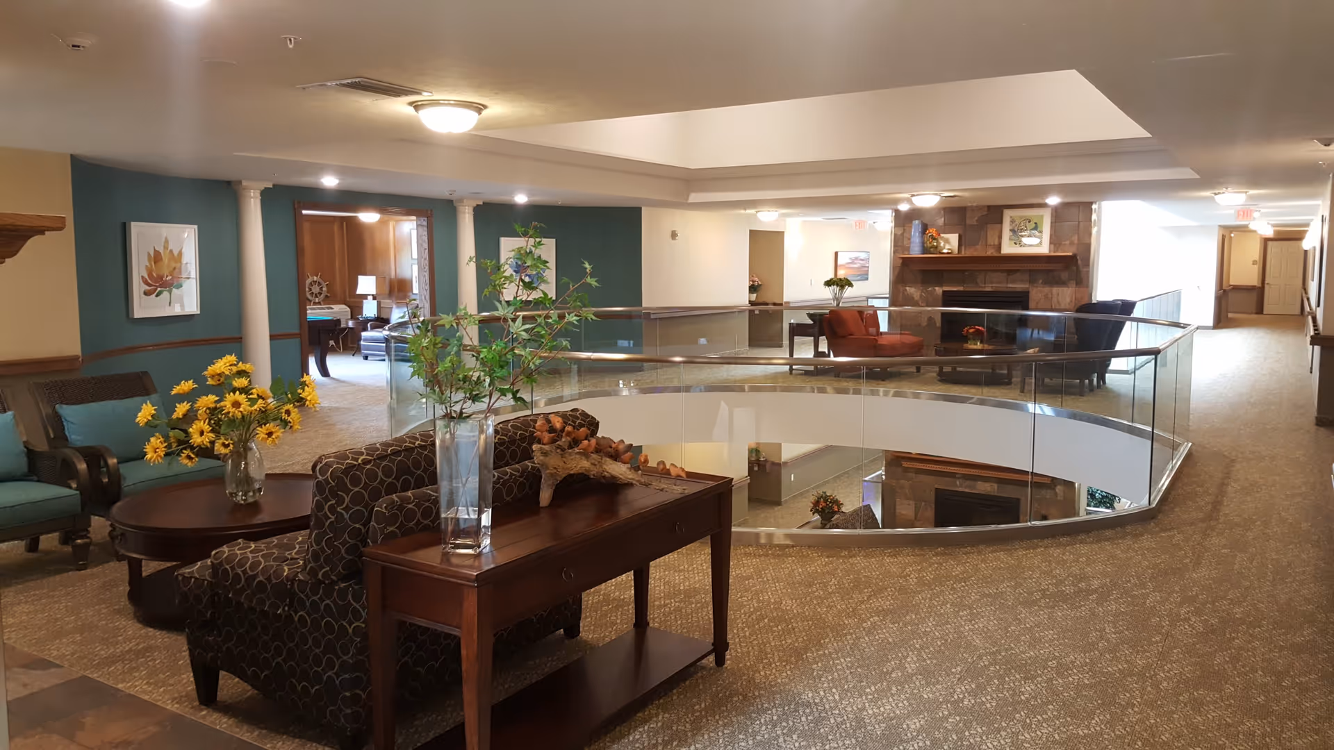 Interior view of a senior living facility lounge area with comfortable seating, a glass railing overlooking a lower level, a fireplace with artwork above it, and decorative plants and flowers on tables. The space is well-lit with ceiling lights and has carpeted floors.