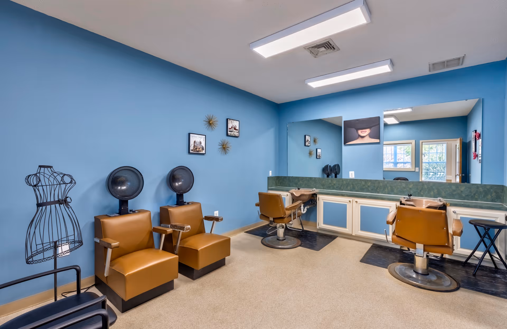 Interior of a hair salon room with blue walls, two brown leather hair drying chairs on the left, two brown salon chairs in front of a large mirror on the right, a black wire dress form, and framed pictures on the wall.