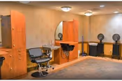 Interior view of a salon area in a nursing home with wooden cabinets, a black salon chair, a wall-mounted mirror, a black sink, and two black fans on stands against the wall.