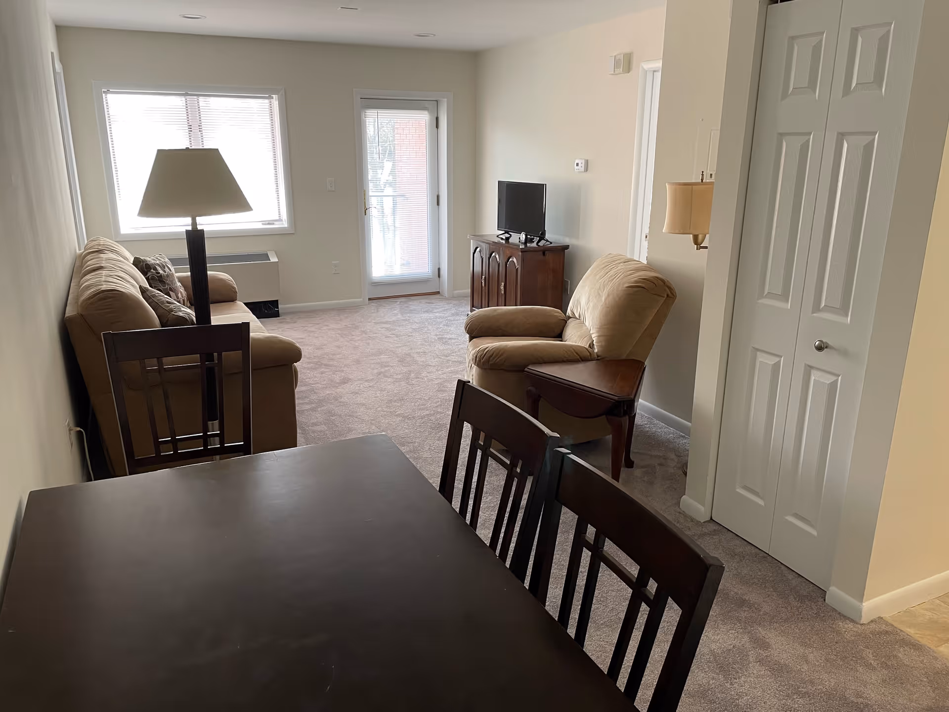 Interior view of a senior living facility's common area featuring a dining table with chairs in the foreground, a beige sofa and armchair, a floor lamp, a small TV on a wooden cabinet, and a door with a window leading outside. The room has carpeted flooring and light-colored walls.