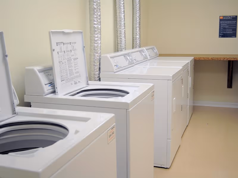 A laundry room with multiple white top-loading washing machines and dryers lined up against a beige wall. There are silver ventilation ducts above the machines and a wooden folding table attached to the wall in the background.