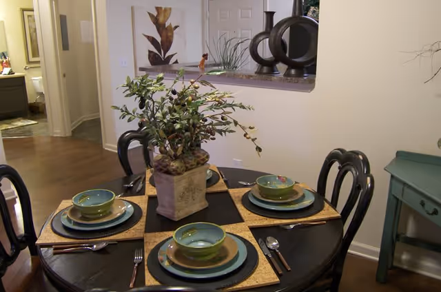 A round dining table set for four with green and blue dishware, silverware, and woven placemats. A decorative plant centerpiece is in the middle of the table. The room has wooden flooring and light-colored walls, with a small green side table and decorative items on a ledge in the background.