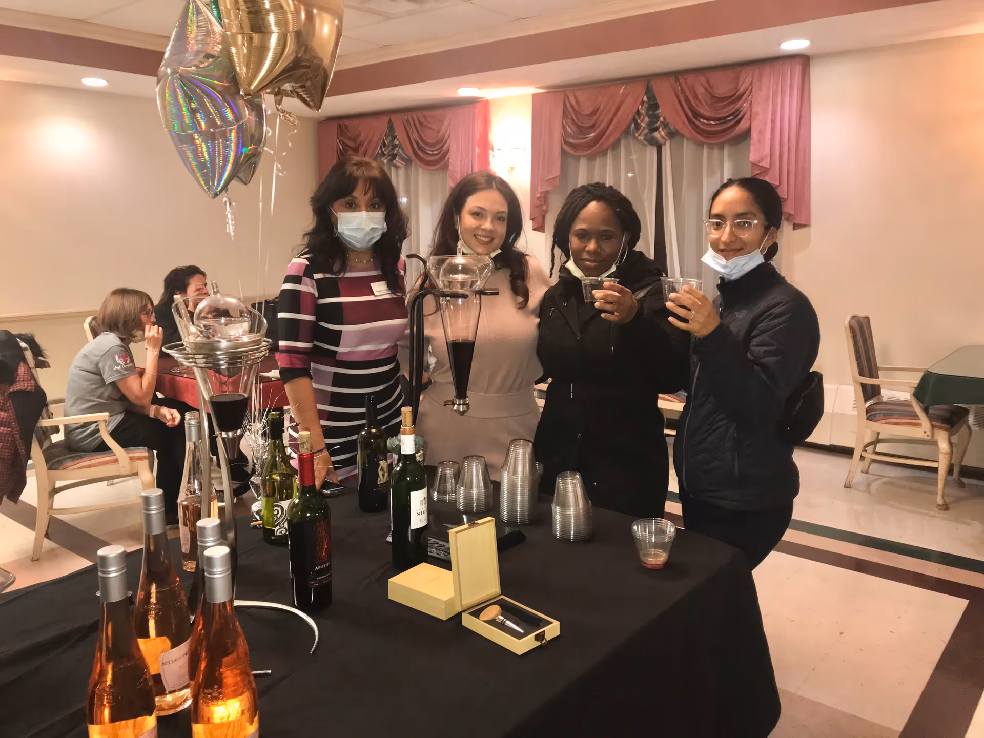 Four women standing behind a table with bottles of wine, plastic cups, and a wine dispenser in a room with pink curtains and balloons. Two women are wearing face masks, and two are holding drinks. In the background, two other women are seated and engaged in conversation.