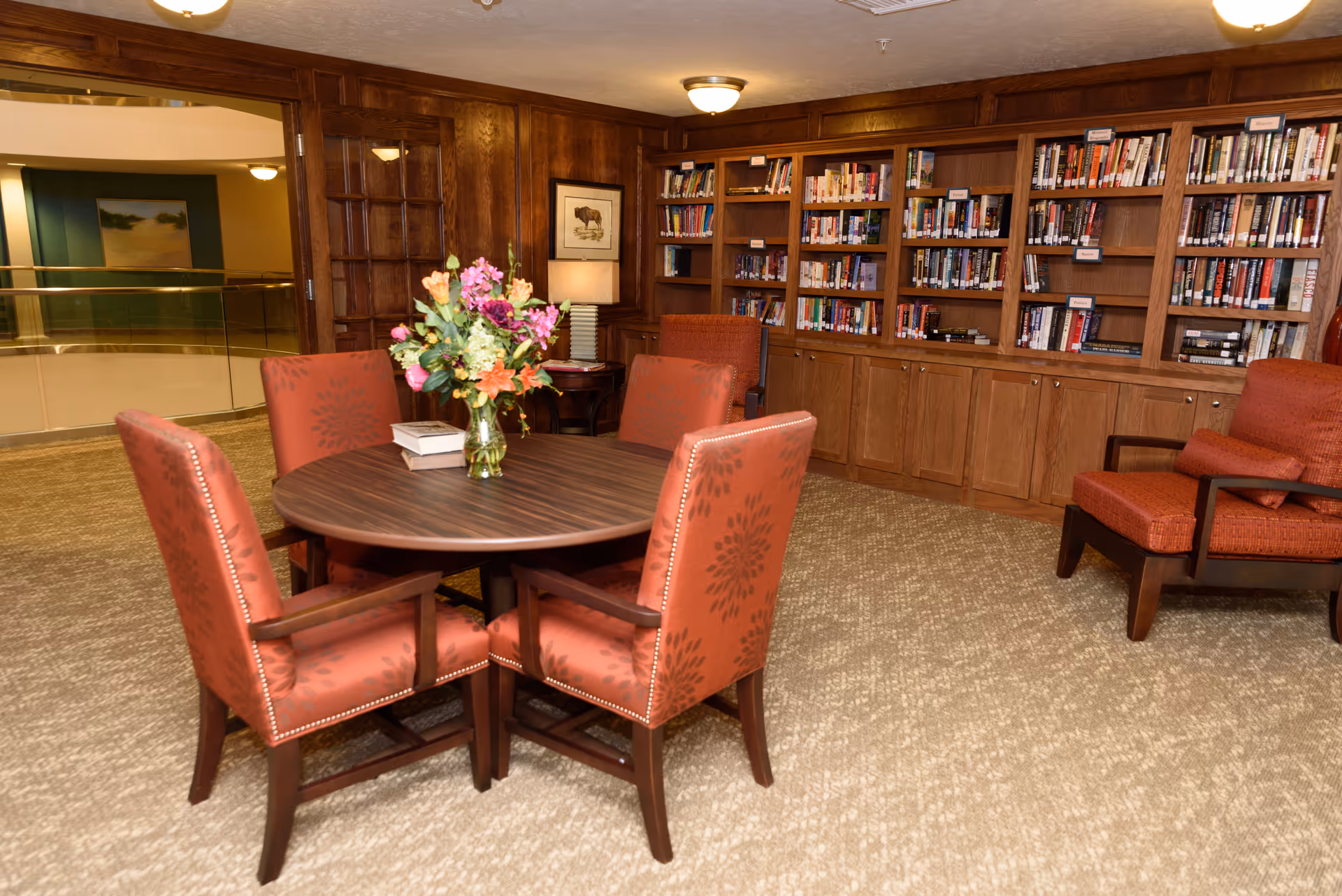 A cozy library room with wooden bookshelves filled with books along the back wall. In the center, there is a round wooden table with a vase of colorful flowers and two books on it, surrounded by four red upholstered chairs. To the right, there is a matching red armchair with a cushion. The room has carpeted flooring and warm lighting.