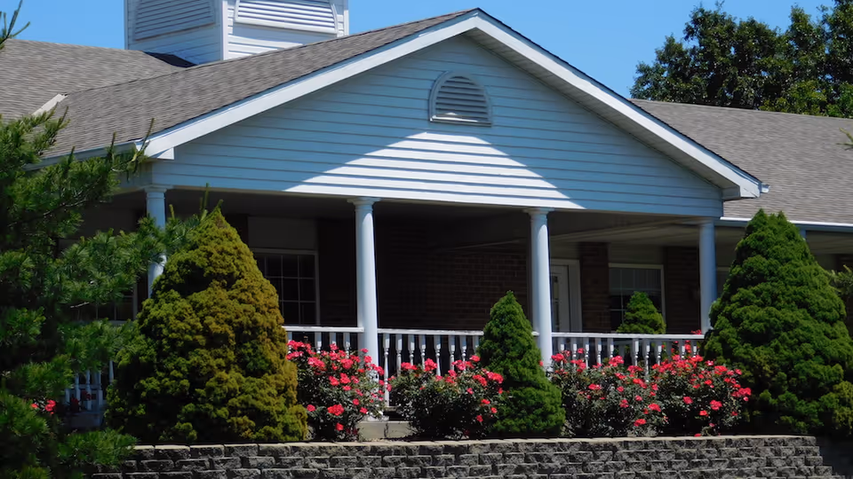 Front porch of a single-story brick building with white columns and railing, flanked by trimmed evergreens and blooming pink rose bushes under a blue sky.