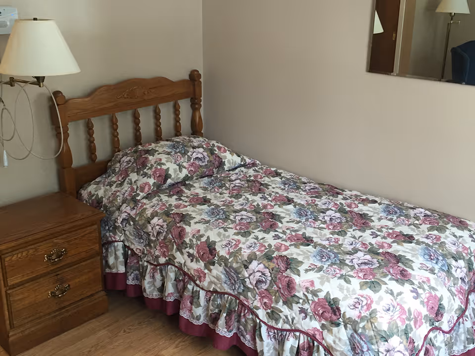 Single bed with a floral bedspread and wooden headboard next to a wooden nightstand and wall lamp in a small bedroom.