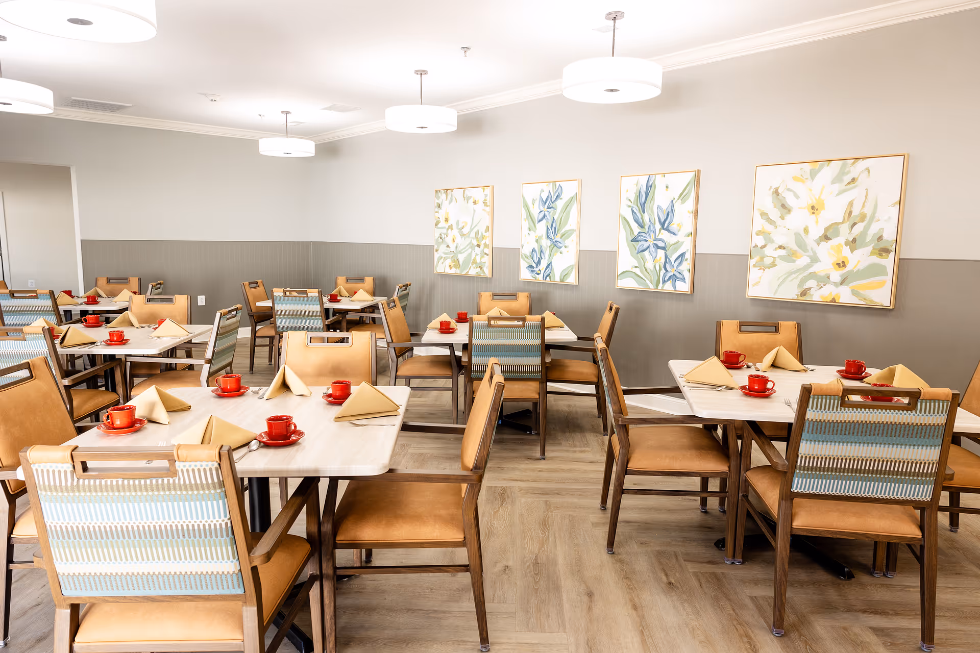 A bright dining room with multiple tables and chairs arranged neatly. Each table is set with red cups and saucers, beige folded napkins, and silverware. The walls are painted in two tones with floral paintings hanging on the upper half. Modern round ceiling lights illuminate the room, and the floor has a light wood finish.