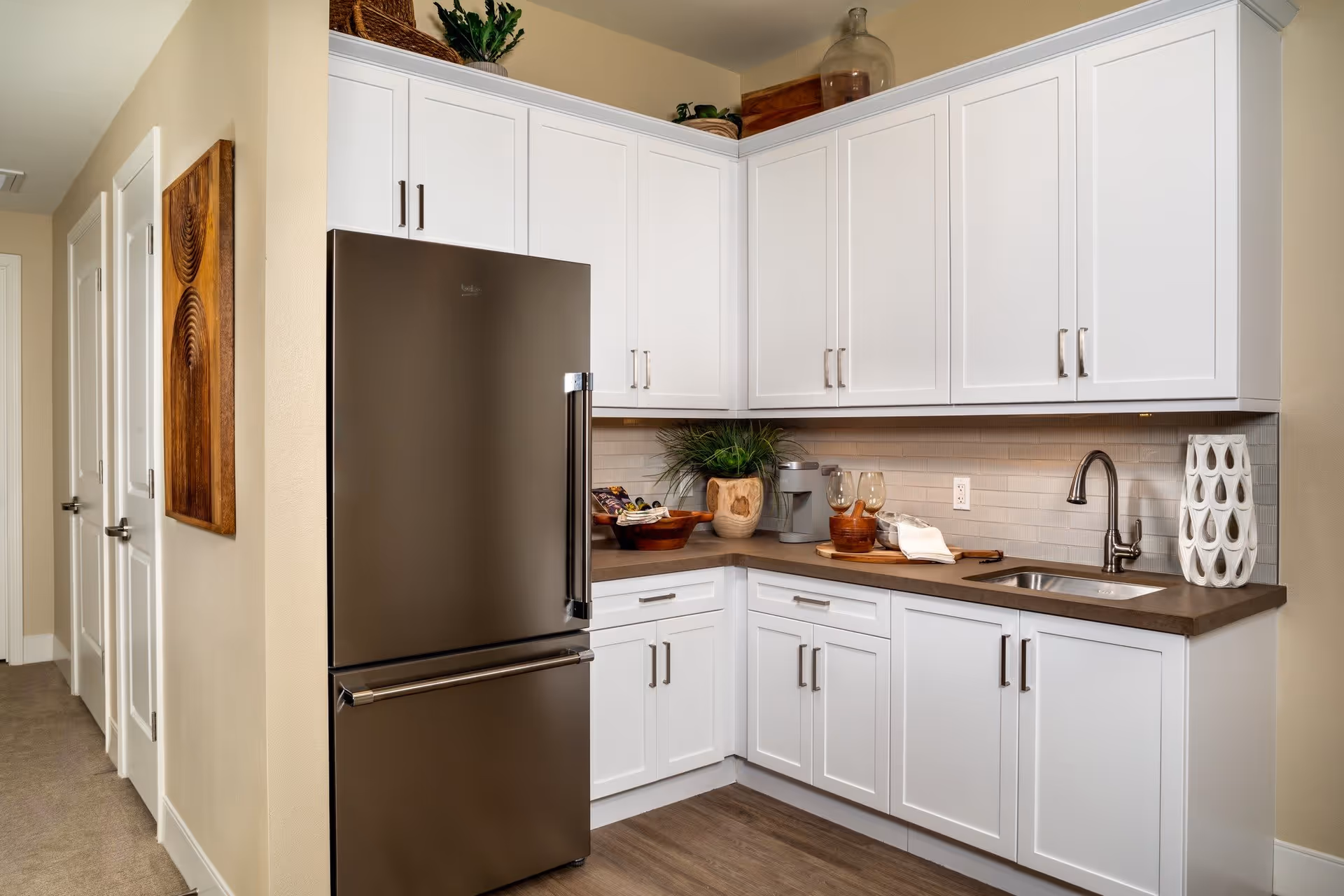 Modern kitchen corner with white cabinets, a stainless steel refrigerator, a sink with a faucet, and decorative items including a plant, glassware, and a ceramic vase on the countertop.