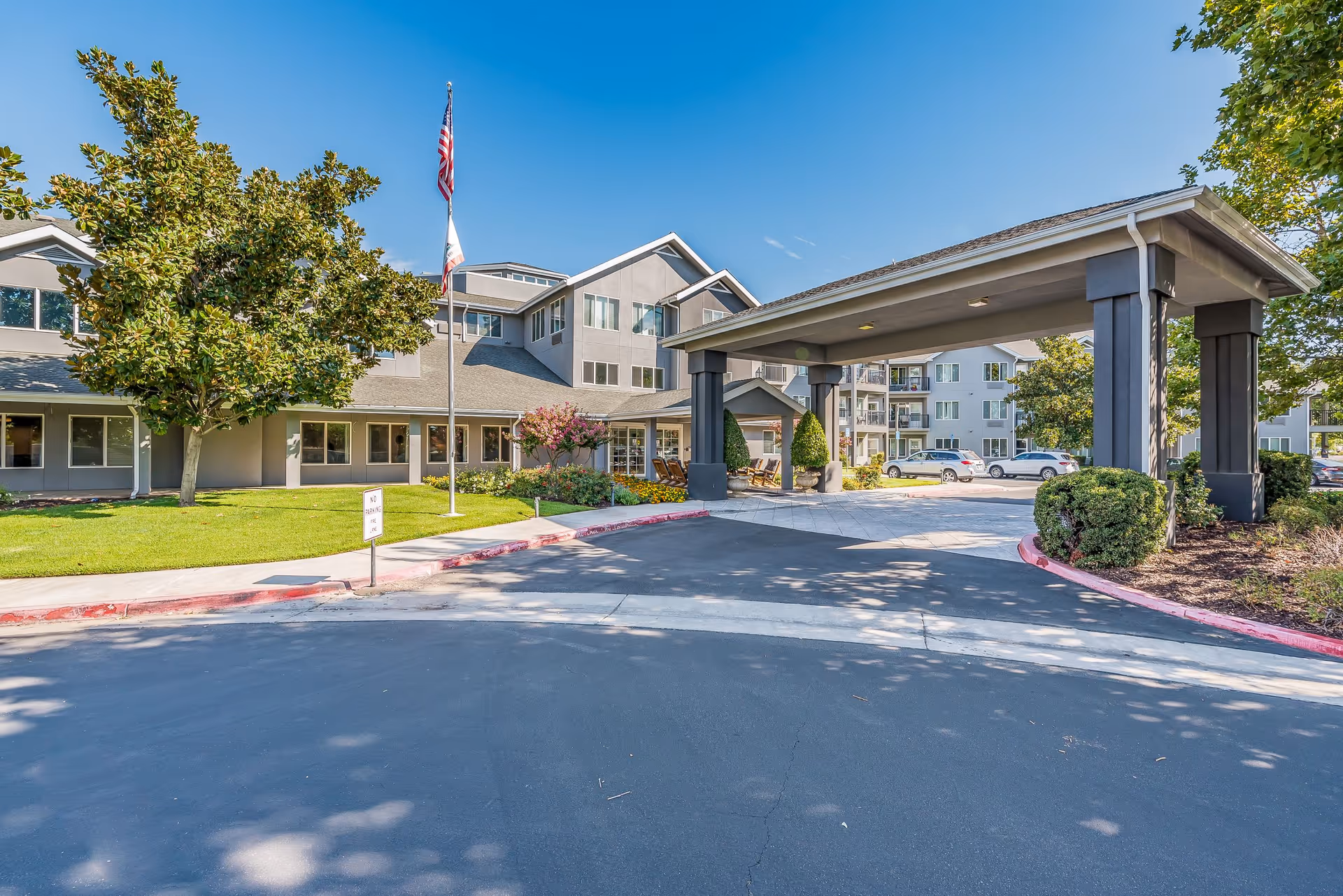 Exterior view of Solstice Senior Living at Clovis showing the main entrance with a covered drop-off area, a flagpole with American and state flags, surrounding greenery including trees and bushes, and a clear blue sky.