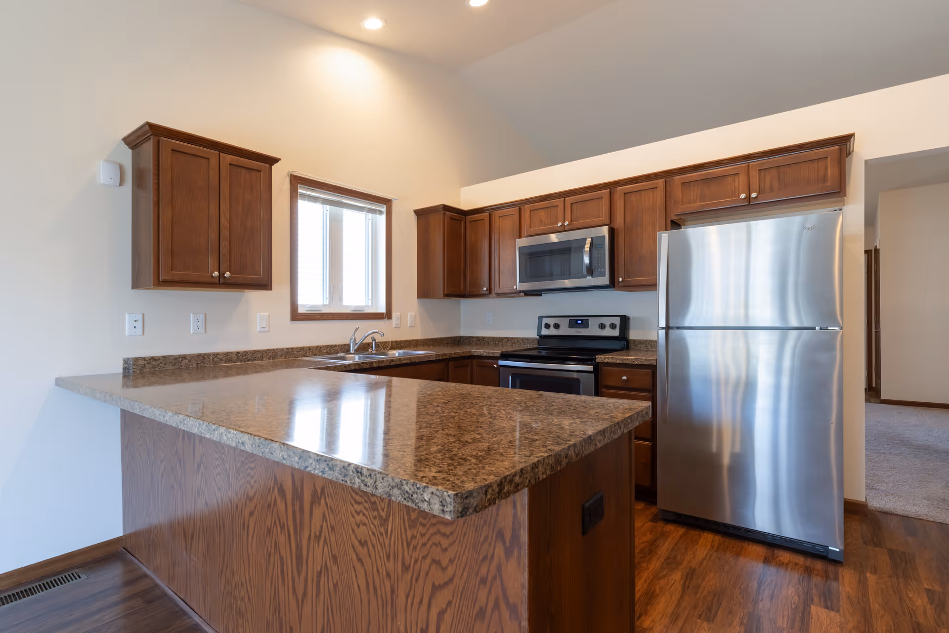 A modern kitchen with wooden cabinets, a granite countertop island, stainless steel refrigerator, stove, and microwave. There is a window above the sink allowing natural light into the space.