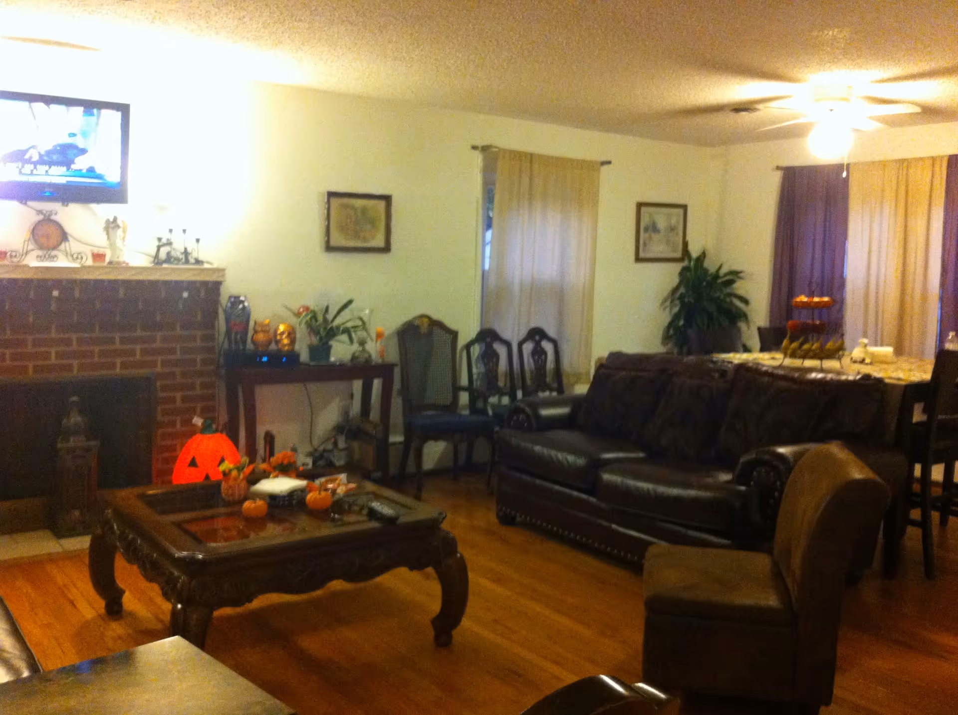 A cozy living room with a dark leather sofa, a wooden coffee table decorated with small pumpkins and flowers, a brick fireplace with a TV mounted above it, and a dining area with a table and chairs in the background. The room has hardwood floors, framed pictures on the walls, and curtains on the windows.