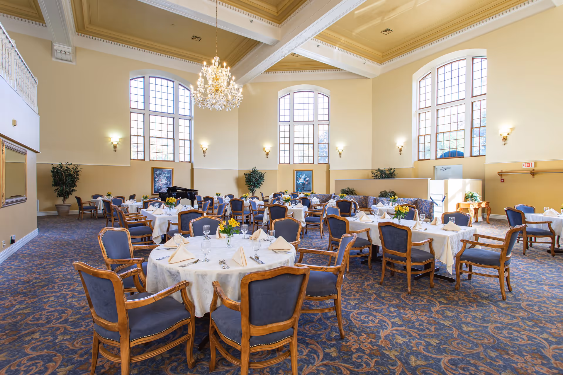 Sunlit formal dining room with round tables set with white linens and wooden chairs under large arched windows and a chandelier.