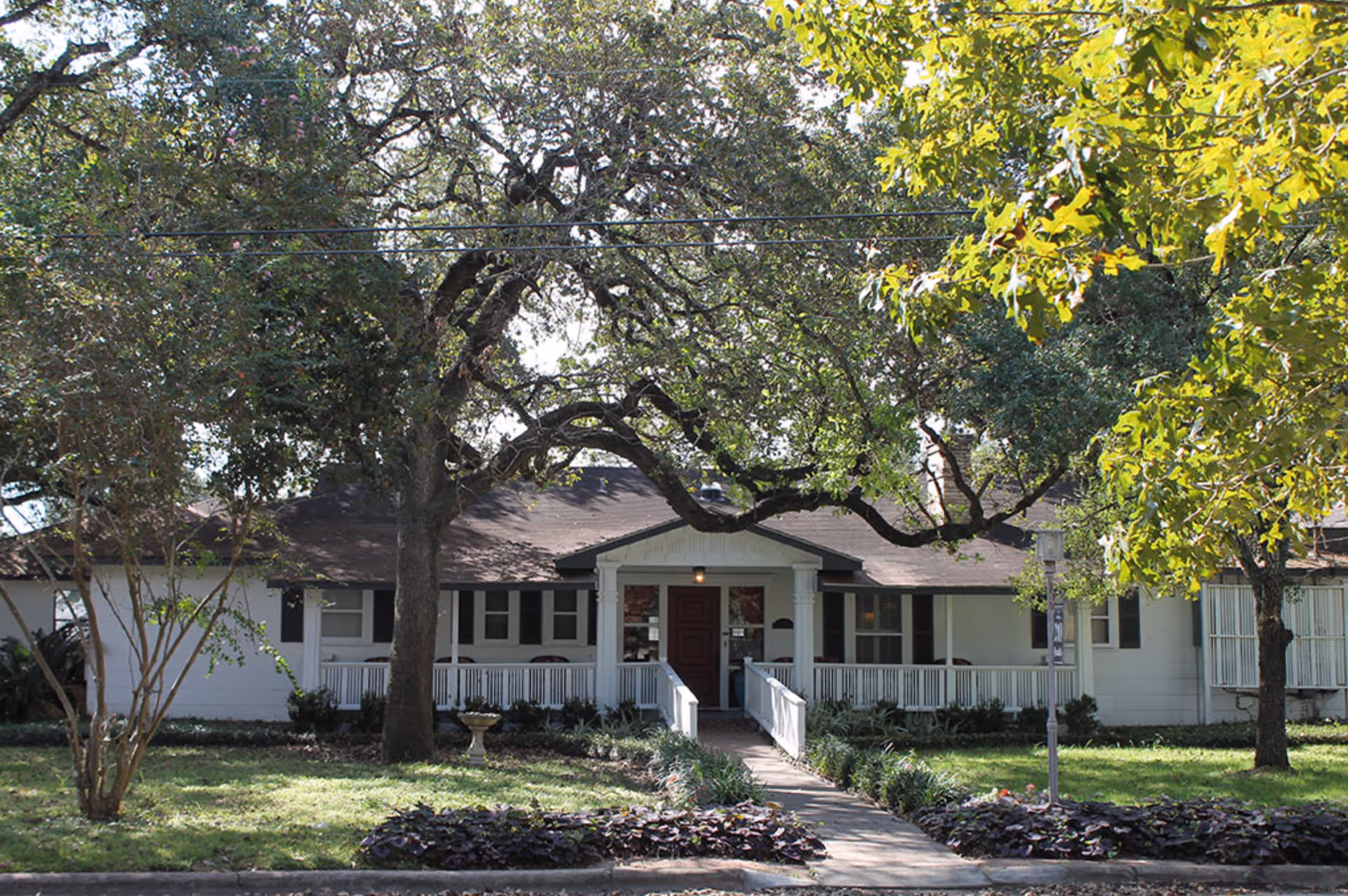 Front exterior view of Romberg House Assisted Living, a single-story building with a dark roof and white walls. The entrance has a small porch with white railings and a ramp leading up to a dark red door. Large trees with green leaves surround the building, casting shadows on the lawn and walkway.
