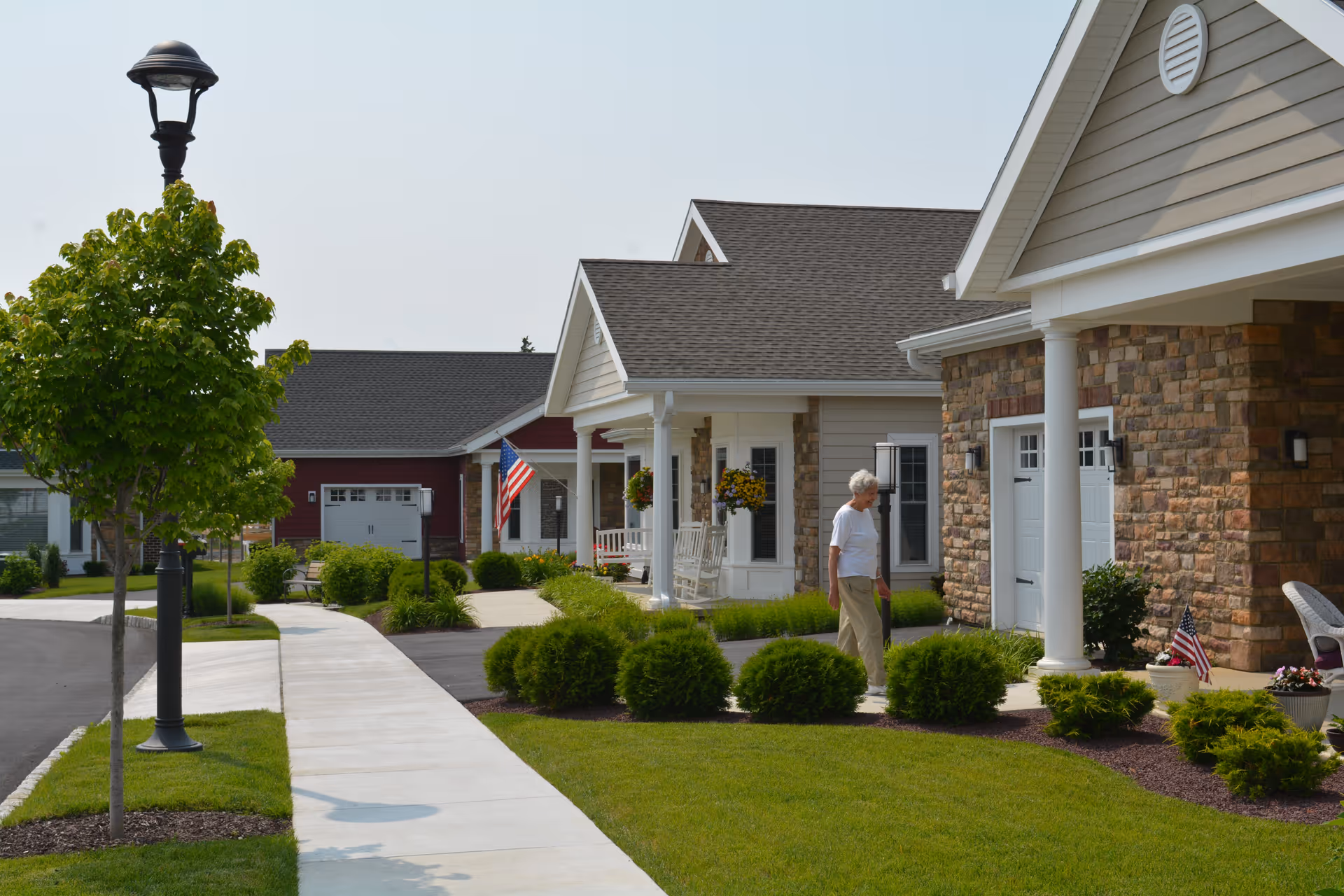 A senior woman walking on a sidewalk in front of single-story residential buildings with stone and siding exteriors, manicured lawns, bushes, and American flags. There is a street lamp and a tree along the sidewalk.