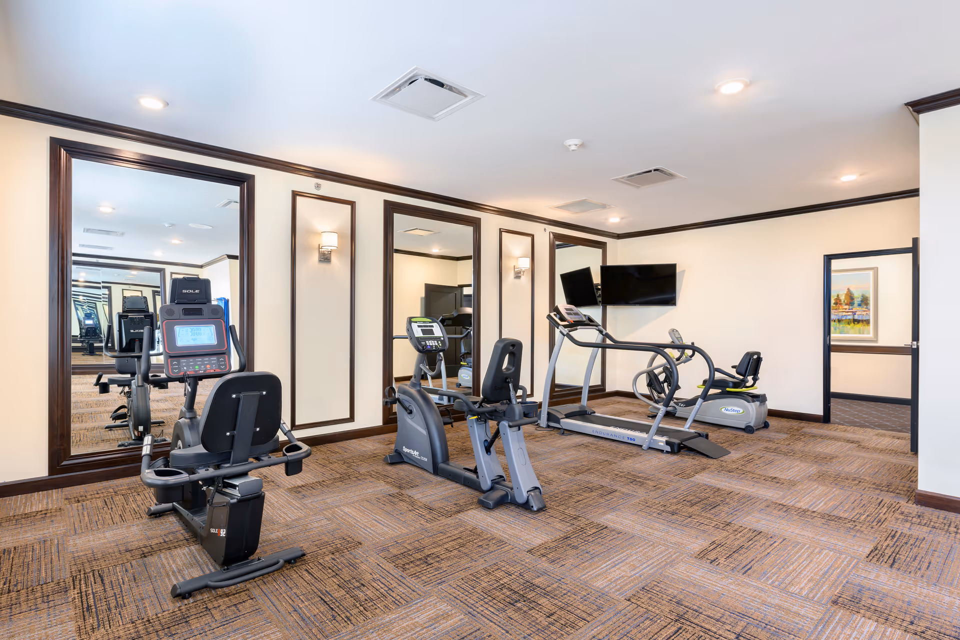 A fitness room with exercise equipment including stationary bikes, a treadmill, and an elliptical machine. The room has large mirrors on the walls, beige walls with dark wood trim, and a wall-mounted TV. The floor is carpeted with a patterned design.