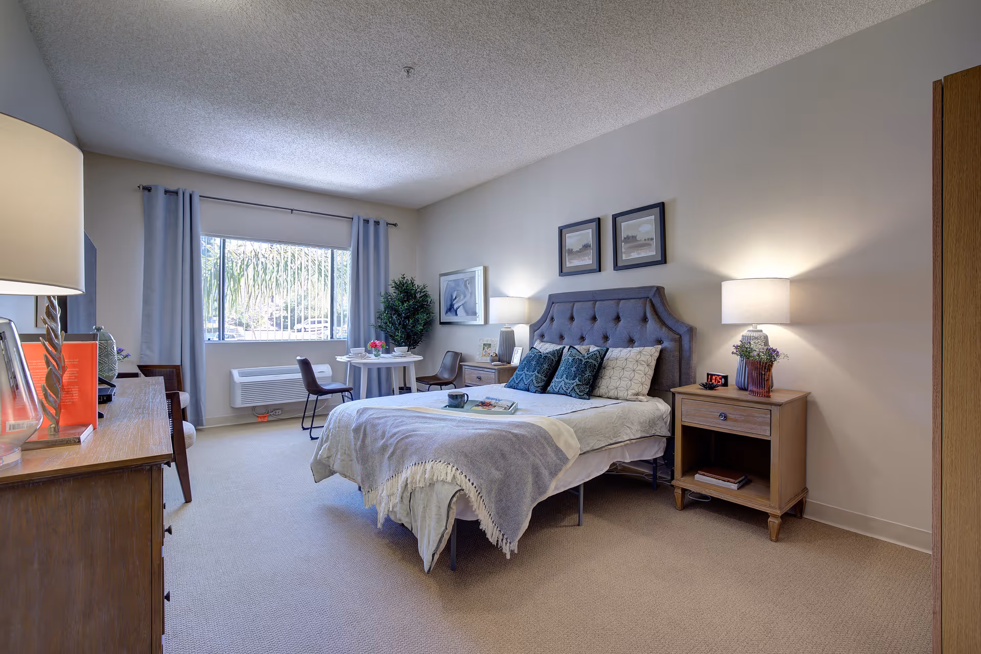 A well-lit senior living bedroom with a large window covered by light gray curtains. The room features a bed with a tufted headboard, blue and white pillows, and a gray throw blanket. There are two wooden nightstands with lamps and decorative items, a small round table with two chairs near the window, a dresser with a lamp and books, and a potted plant in the corner.