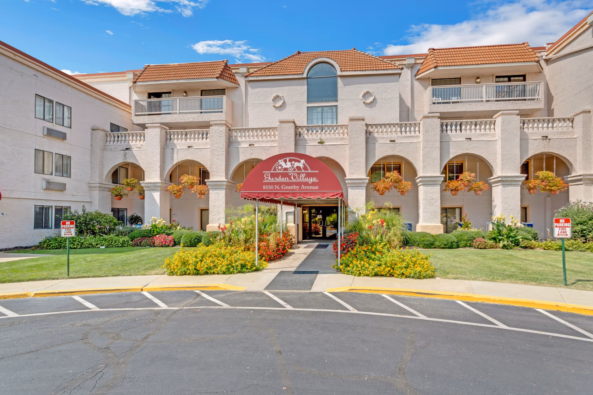 Front exterior view of Garden Village senior living facility with a beige stucco building featuring arches, balconies, and a red awning over the entrance. The entrance is surrounded by well-maintained green lawns and colorful flower beds under a partly cloudy blue sky.