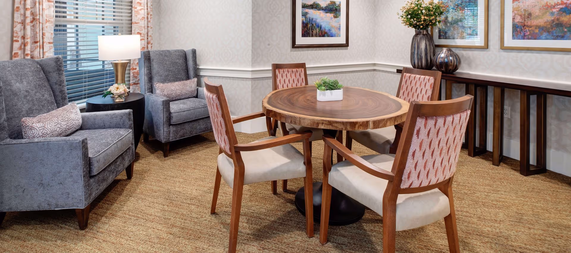A cozy sitting area with a round wooden table surrounded by four wooden chairs with patterned backs and cushioned seats. Two gray upholstered armchairs with decorative pillows are positioned near a window with blinds and floral curtains. A small round side table with a lamp and a flower arrangement is between the armchairs. The walls are decorated with framed artwork and a console table with vases and flowers.