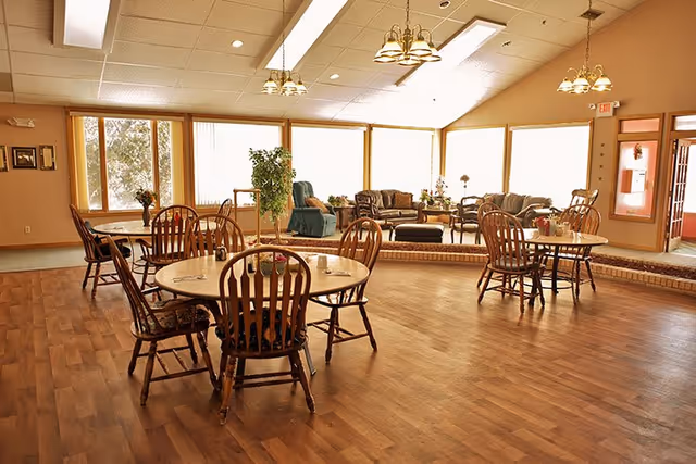 Spacious common dining room with round wooden tables and chairs and a raised lounge area with sofas by large windows.