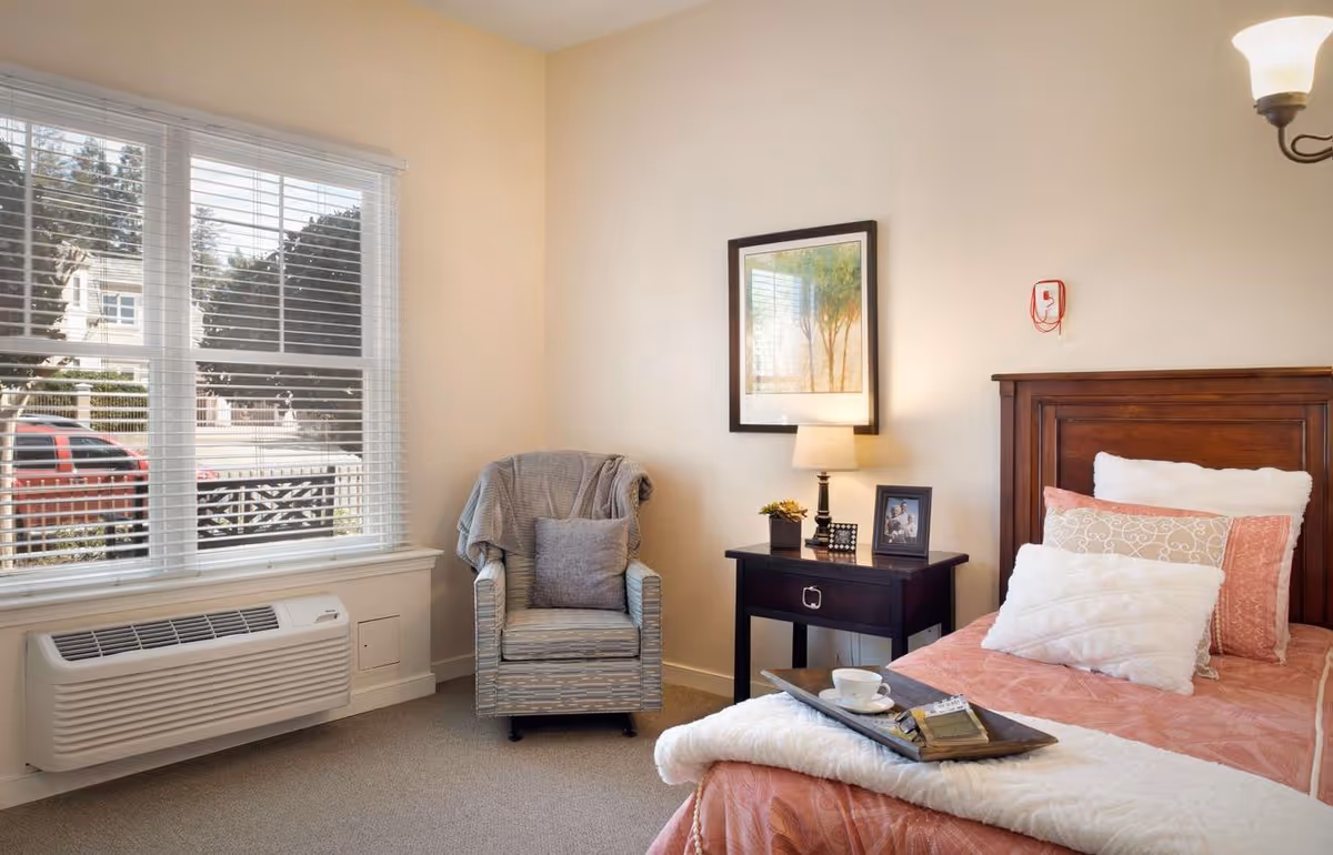A cozy bedroom with a single bed covered in a pink and white comforter and multiple pillows. A wooden nightstand beside the bed holds a lamp, a small plant, and framed photos. Next to the nightstand is a comfortable armchair with a gray throw blanket and pillow. A large window with white blinds lets in natural light, and an air conditioning unit is installed below the window. A framed picture hangs on the wall above the nightstand.