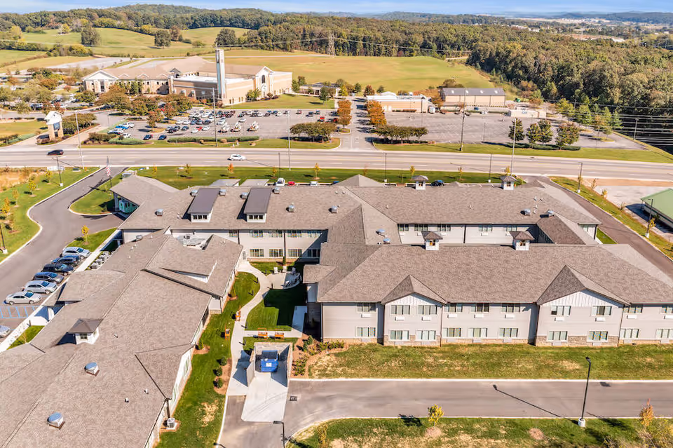 Aerial view of a senior living facility with multiple connected buildings featuring pitched roofs, surrounded by green lawns and parking areas. The facility is located near a road with additional buildings and wooded hills in the background under a clear sky.