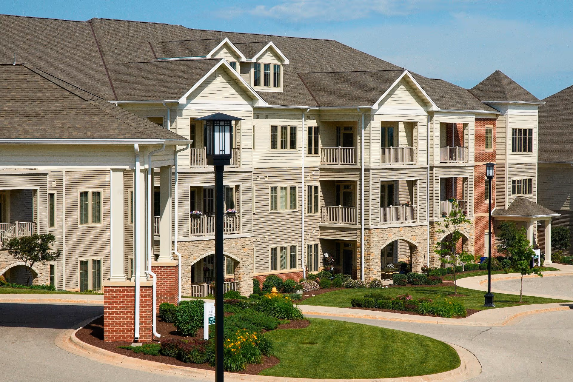 Exterior view of a multi-story senior living facility building with beige siding, brick accents, balconies, and a covered entrance. The surrounding area includes well-maintained landscaping with green grass, shrubs, and trees under a clear blue sky.