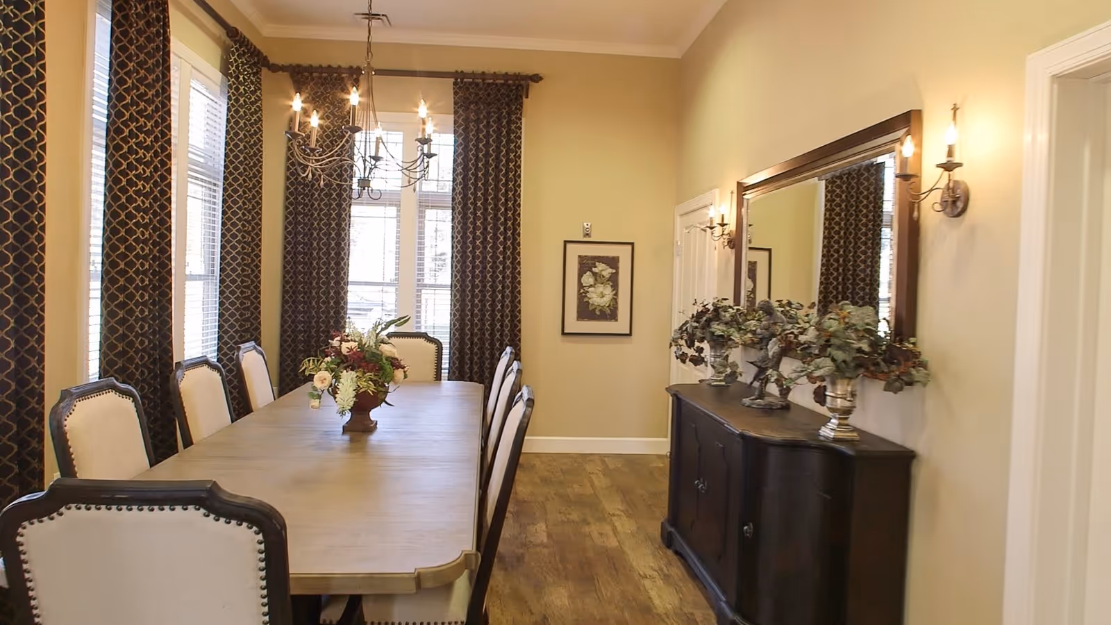 A formal dining room with a long wooden table surrounded by upholstered chairs. The room features large windows with patterned dark curtains, a chandelier hanging above the table, a sideboard with decorative plants and a large mirror on the wall, and warm yellow walls with wooden flooring.