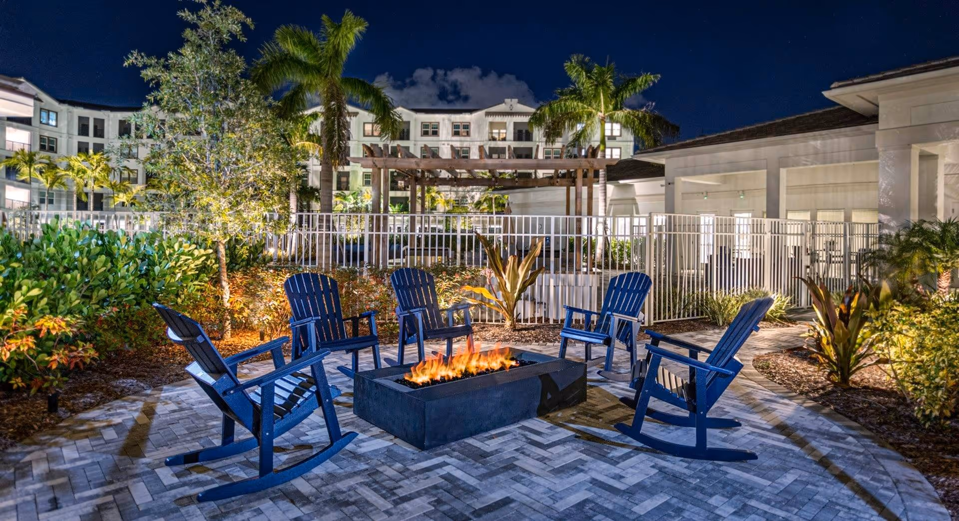 Outdoor seating area at night with six blue rocking chairs arranged around a rectangular fire pit. The area is paved with patterned bricks and surrounded by plants and palm trees. In the background, a multi-story building and a pergola are visible under a dark sky.