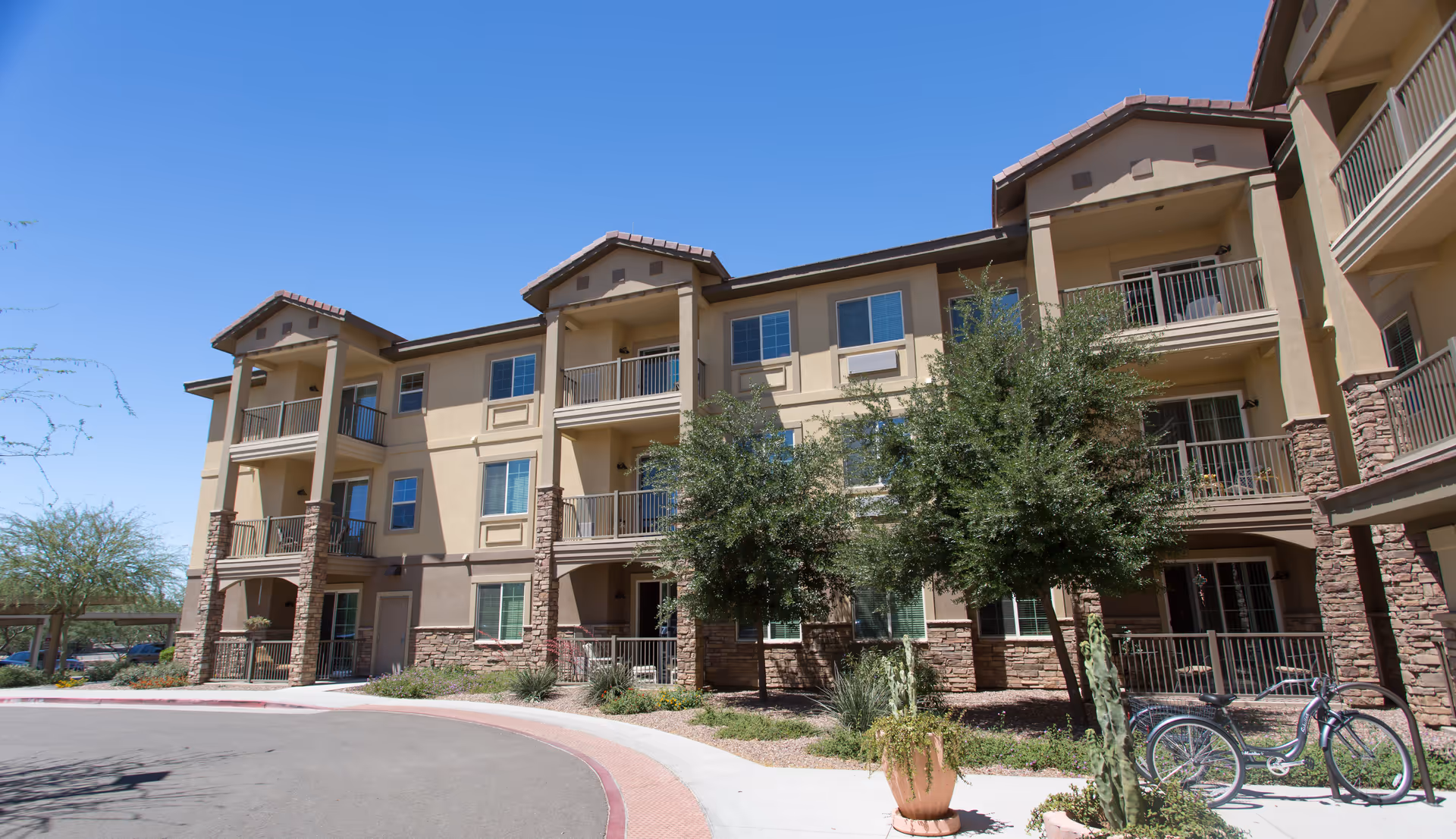 Three-story beige senior living building with balconies, landscaping, and a bicycle near the entrance under a clear blue sky.
