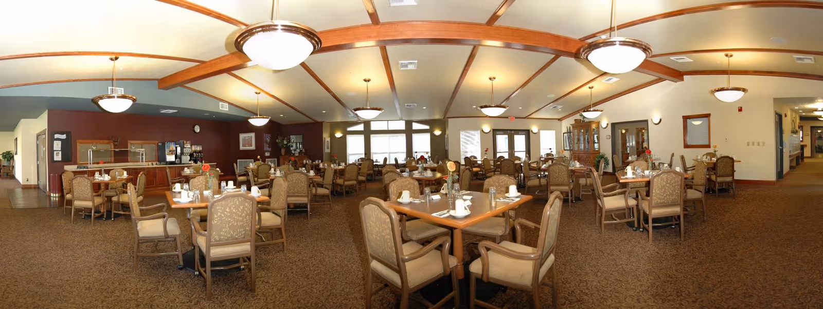 A spacious dining room with multiple wooden tables and cushioned chairs arranged neatly. Each table is set with white cups, saucers, and napkins. The room has a warm color palette with beige walls, carpeted floors, and wooden ceiling beams. Several ceiling lights provide illumination, and large windows at the far end allow natural light to enter. There is a counter area with a coffee machine and other amenities on the left side of the room.