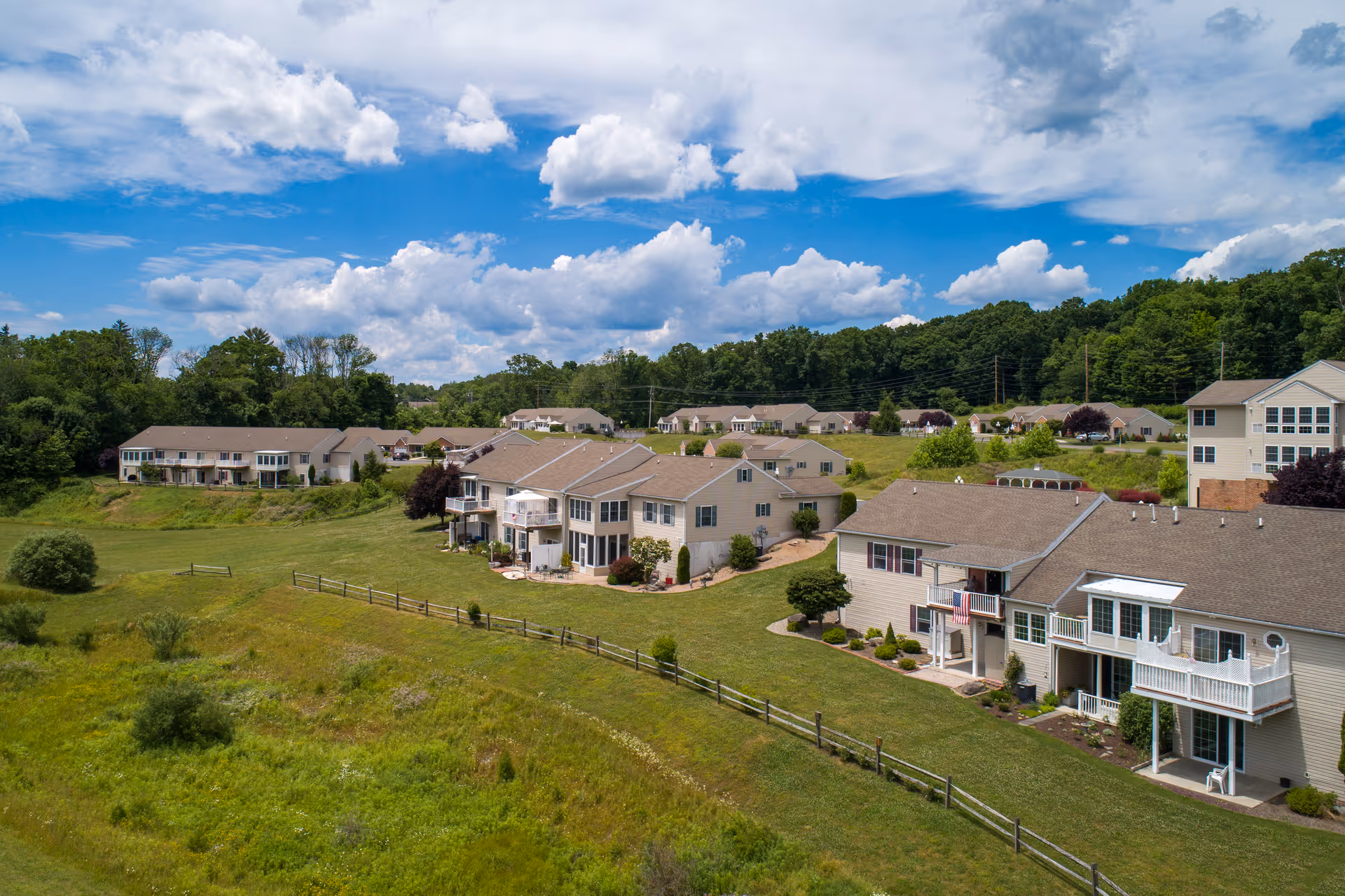 Aerial view of a senior living community with rows of multi-unit buildings, green lawns, and a wooden fence under a partly cloudy blue sky.