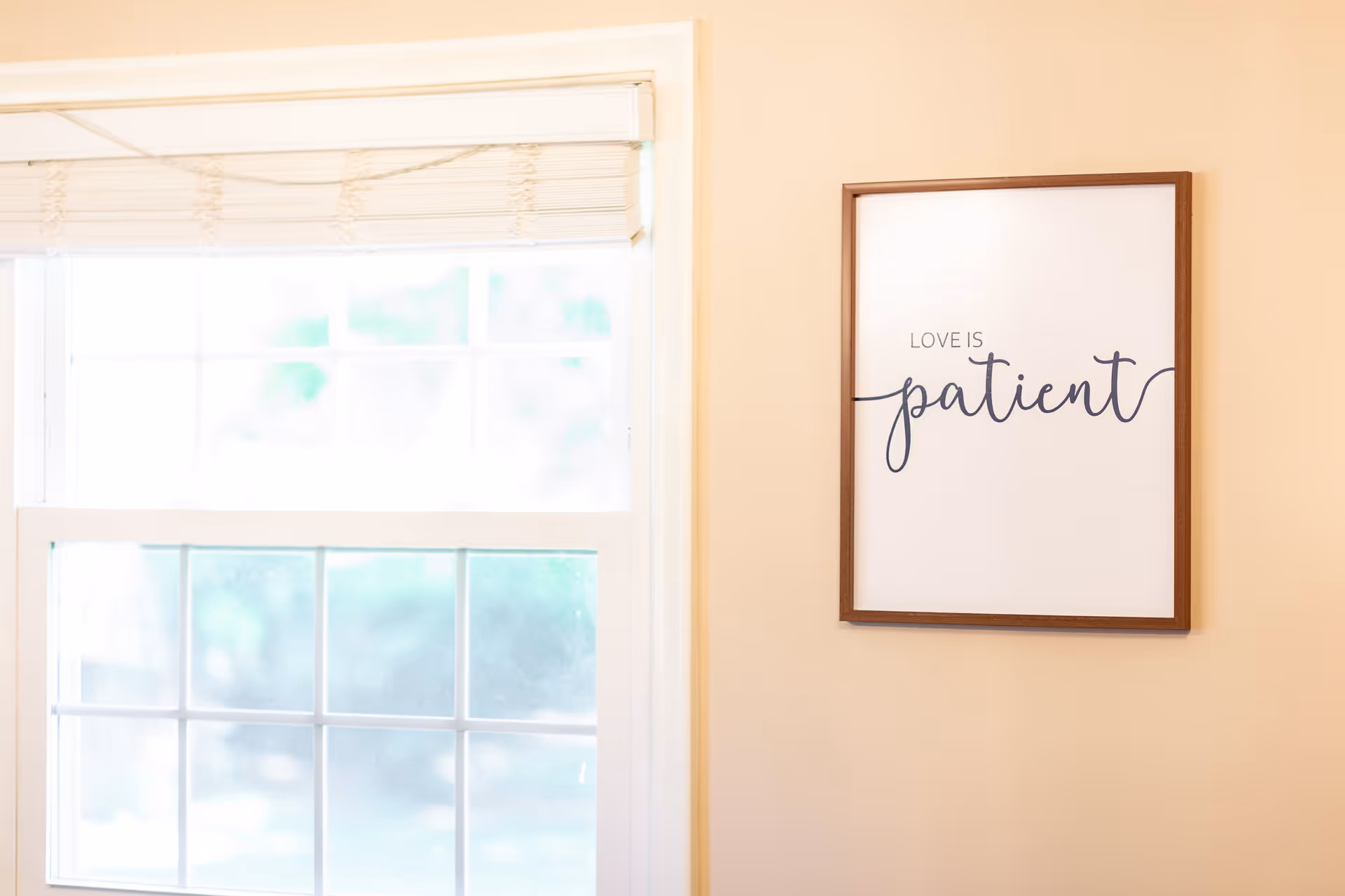 A framed wall art with the phrase 'Love is patient' written in elegant script hangs on a beige wall next to a window with white blinds partially drawn.