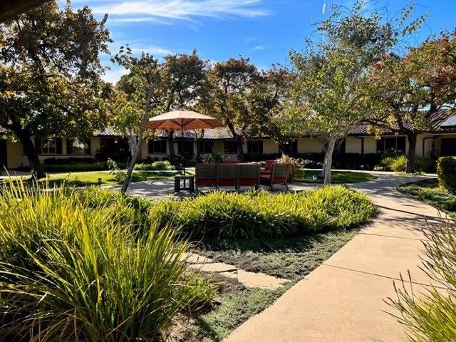 Outdoor garden area at Wood Glen Senior Living with a circular seating arrangement under a large umbrella surrounded by trees and greenery, with a paved walkway leading through the landscaped space.