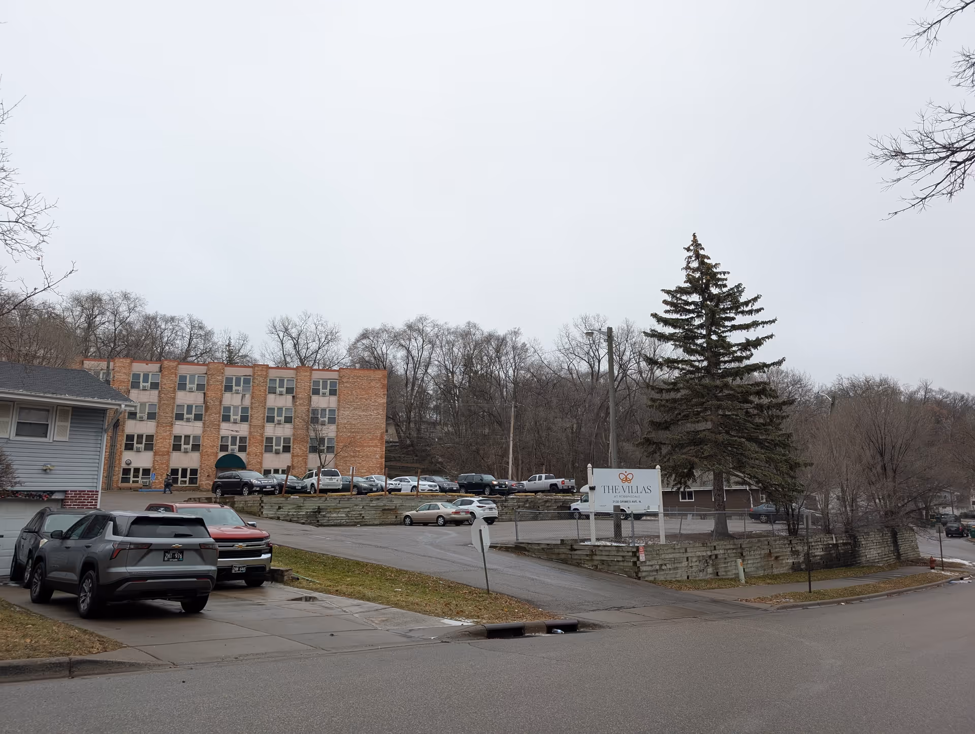 Exterior view of The Villas at Robbinsdale facility showing a multi-story brick building, a parking lot with several cars, a large evergreen tree, and a sign with the facility's name near the entrance.