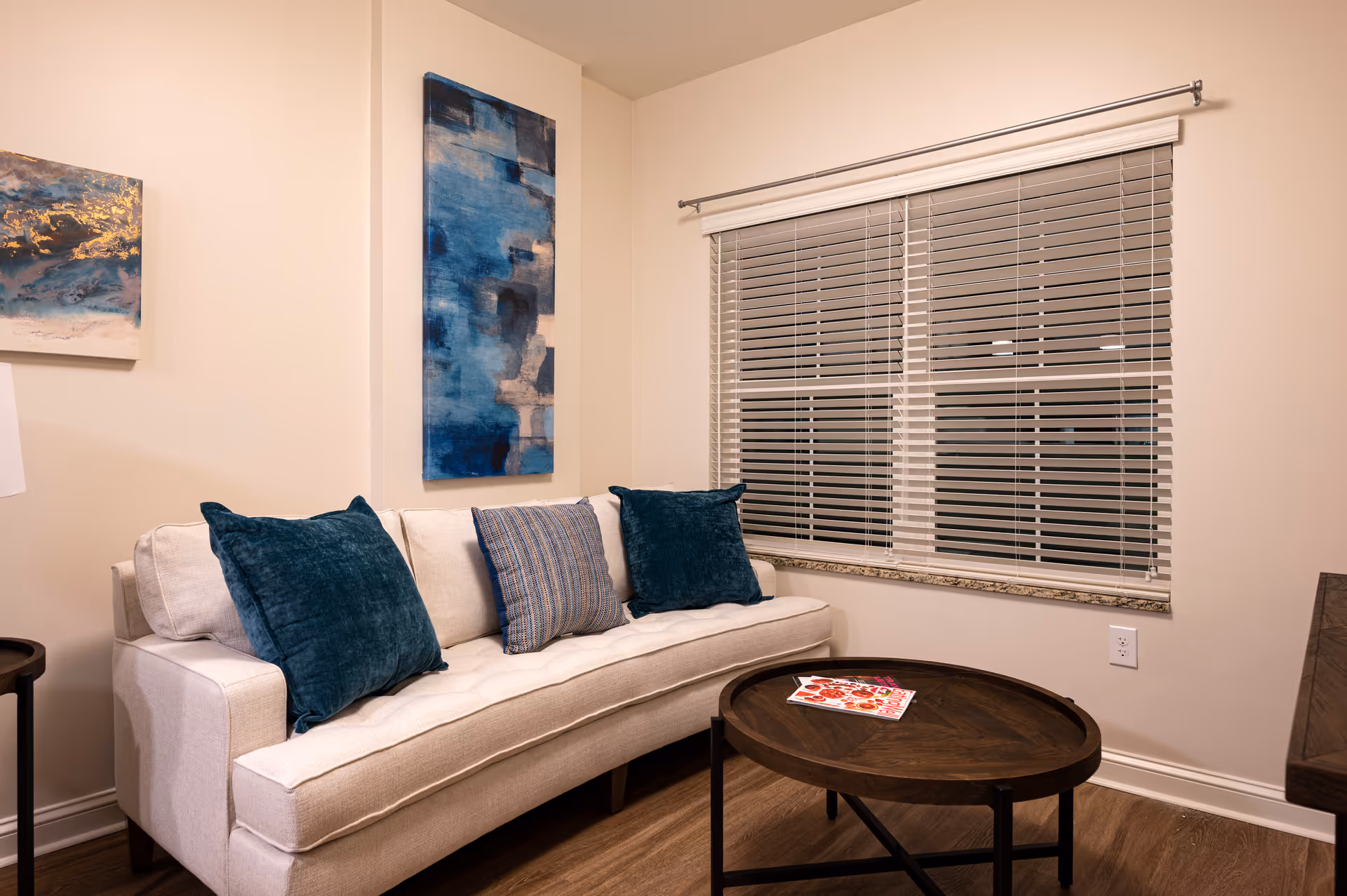 A cozy living room area with a beige sofa adorned with three cushions in blue and striped patterns. There are two abstract blue paintings on the cream-colored walls. A round wooden coffee table with a magazine on top is placed in front of the sofa. A window with closed white blinds is on the right side of the room.