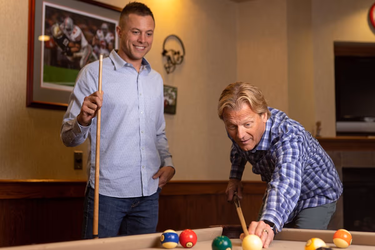 Two men playing pool indoors. One man is leaning over the pool table aiming to strike the cue ball with a pool cue, while the other man stands nearby holding a pool cue and smiling. The room has framed sports pictures on the wall and warm lighting.