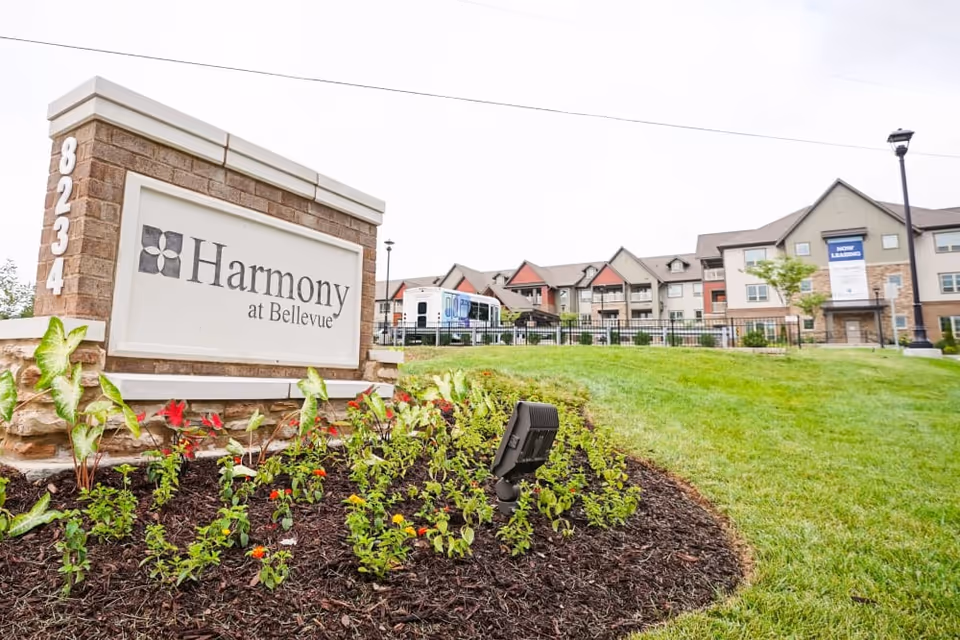 Exterior view of Harmony at Bellevue senior living facility with a large sign displaying the facility name and address 8234. The building is multi-story with a mix of beige and brown tones, surrounded by a well-maintained lawn and flower bed in the foreground.