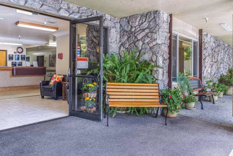 Entrance area of a facility with a glass door open leading inside to a reception desk and seating area. Outside the entrance, there are wooden benches, potted plants, and stone-textured walls.