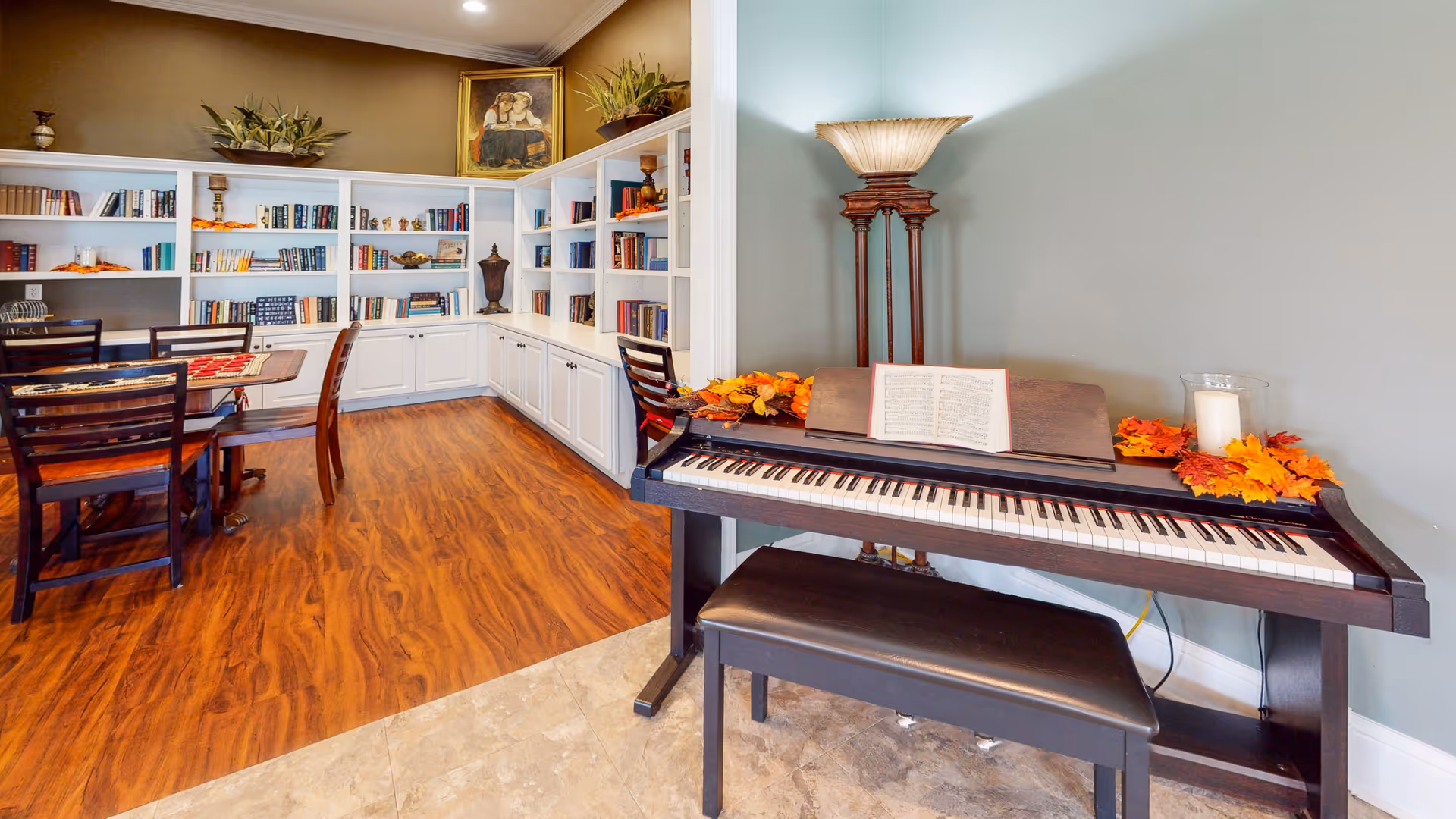 Community lounge with an electronic piano and bench in the foreground and a dining table and built-in bookshelves in the background.