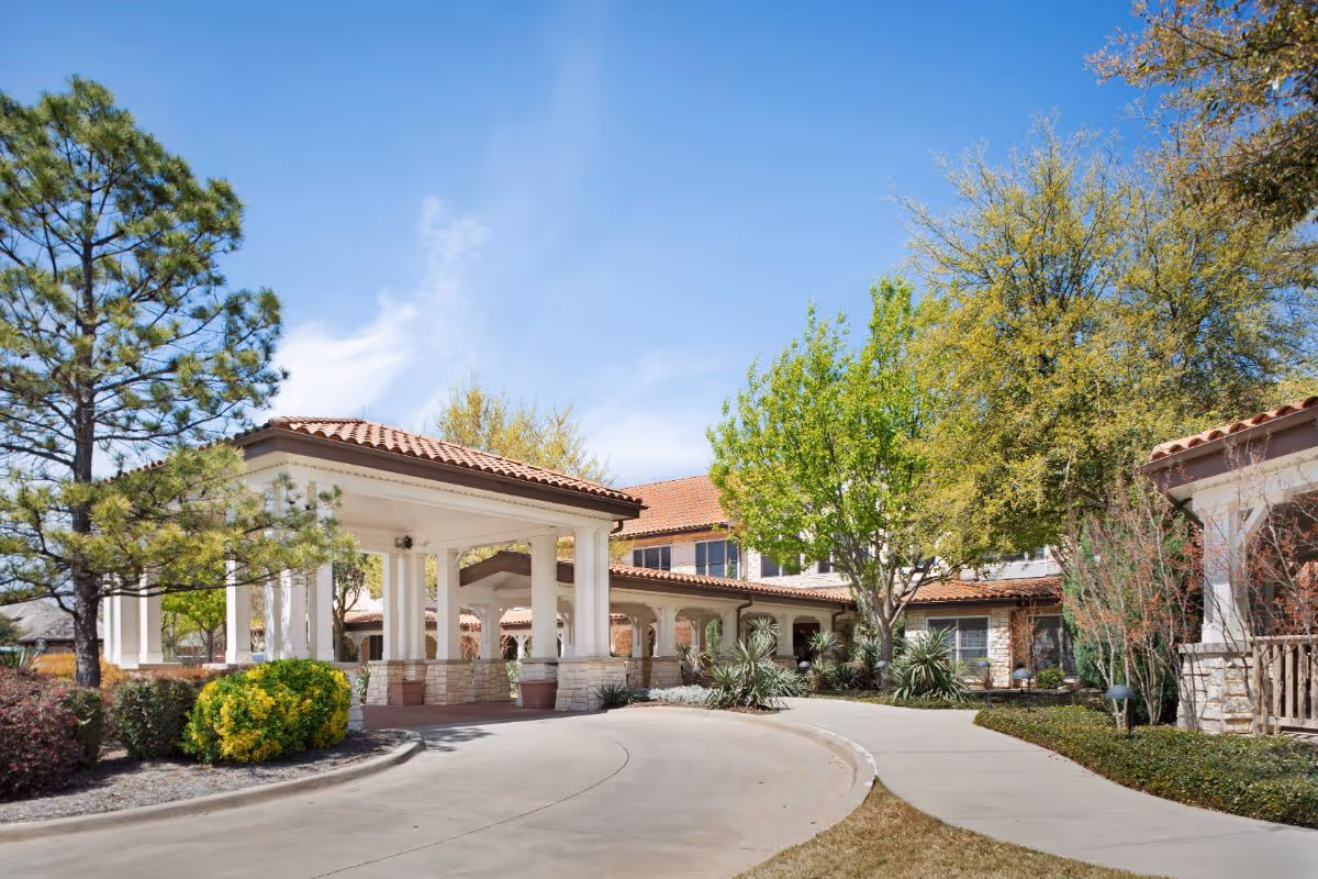 Exterior view of a senior living facility with a covered entrance, surrounded by trees and shrubs under a clear blue sky.