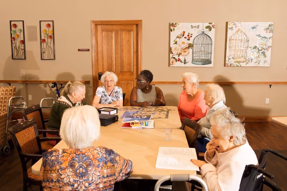 A group of elderly women sitting around a table in a well-lit room, engaging in an activity with puzzles and books. The room has beige walls decorated with floral and birdcage artwork, wooden doors, and chairs. Some women have walkers nearby.